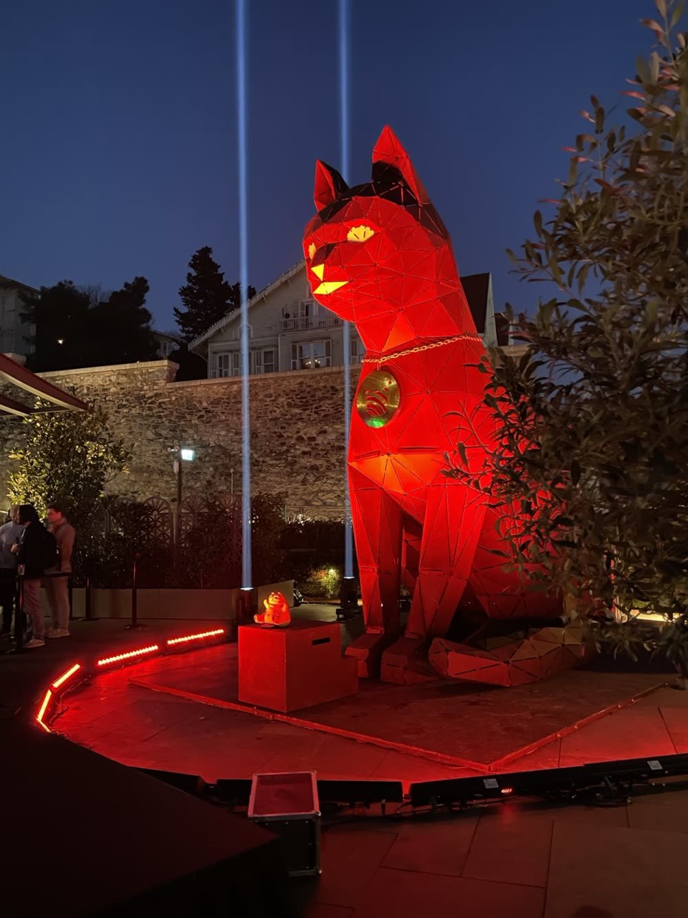 A monumental low-poly metal cat sculpture lit in red at an outdoor evening event