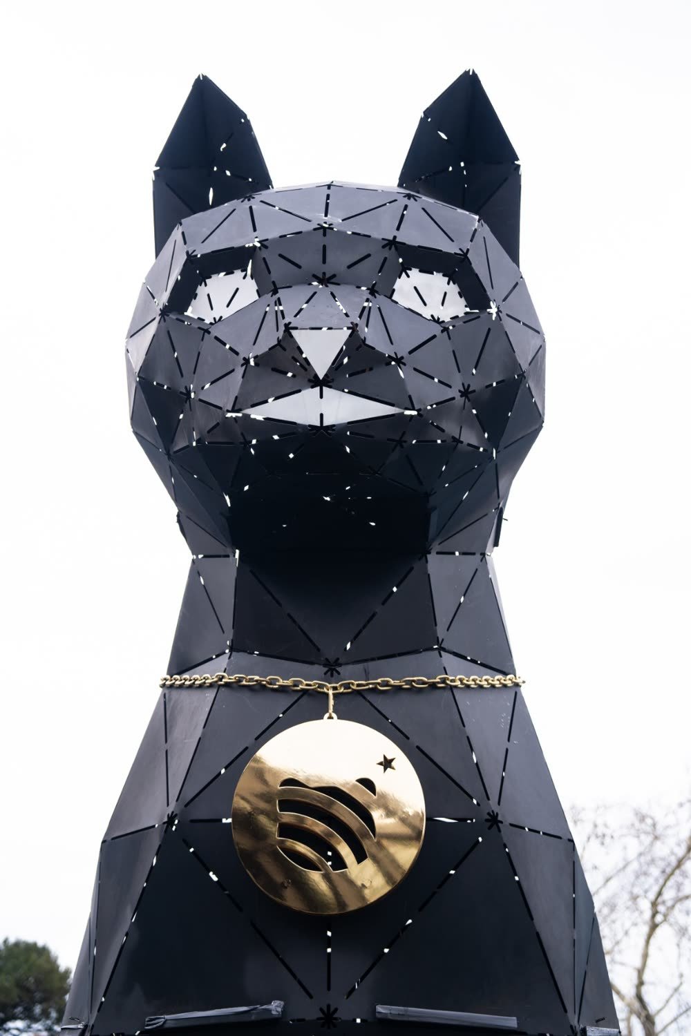 The head of a black low-poly metal cat sculpture with a gold medallion at its neck, in daylight